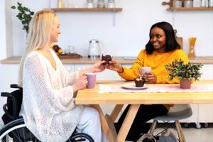 Woman on wheelchair sitting on kithcen dining table with her guardian. Both women are smiling and holding a chocoloate cup cake